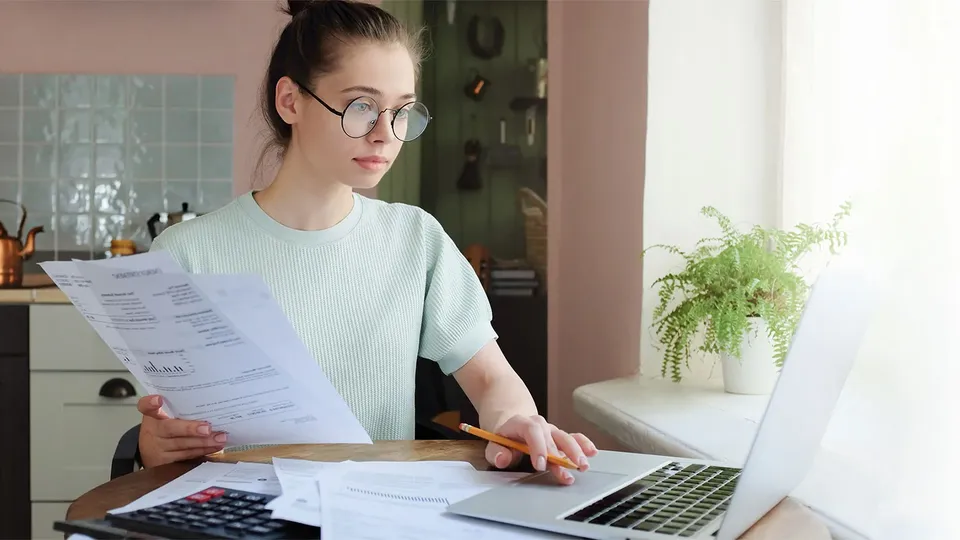 Kvinde gennemgår dokumenter ved skrivebord med bærbar computer og lommeregner.