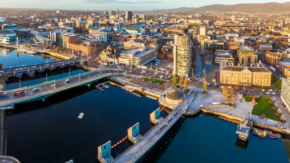 Aerial view of Belfast waterfront with bridges, docks, and city skyline at sunset.