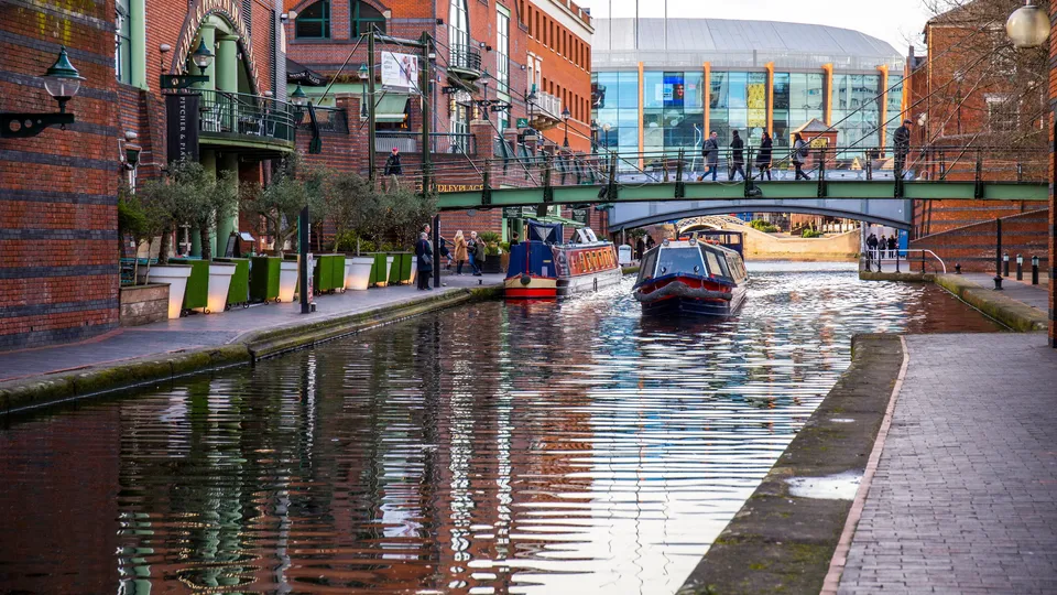 Narrowboats traveling through a canal lock in an urban waterway surrounded by brick buildings and a pedestrian bridge.