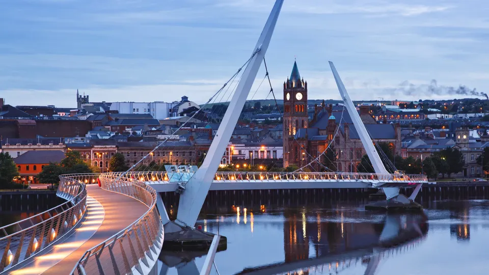 Illuminated pedestrian bridge spanning river with cathedral tower in background at dusk.