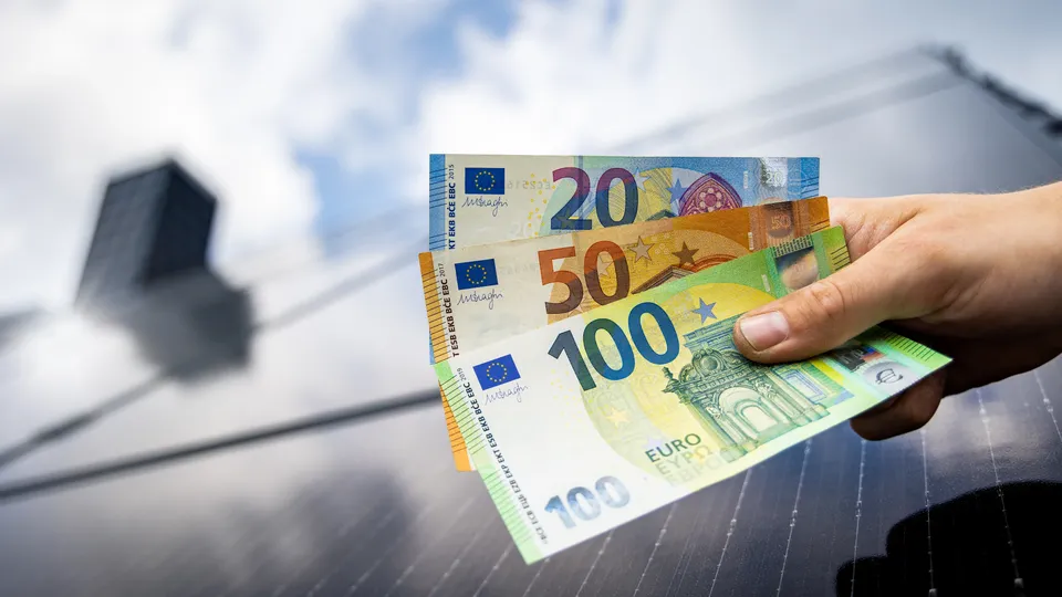 Eine Hand hält mehrere Euro-Banknoten vor einem Solarmodul als Symbolbild für die Einspeisevergütung von Photovoltaikanlagen.