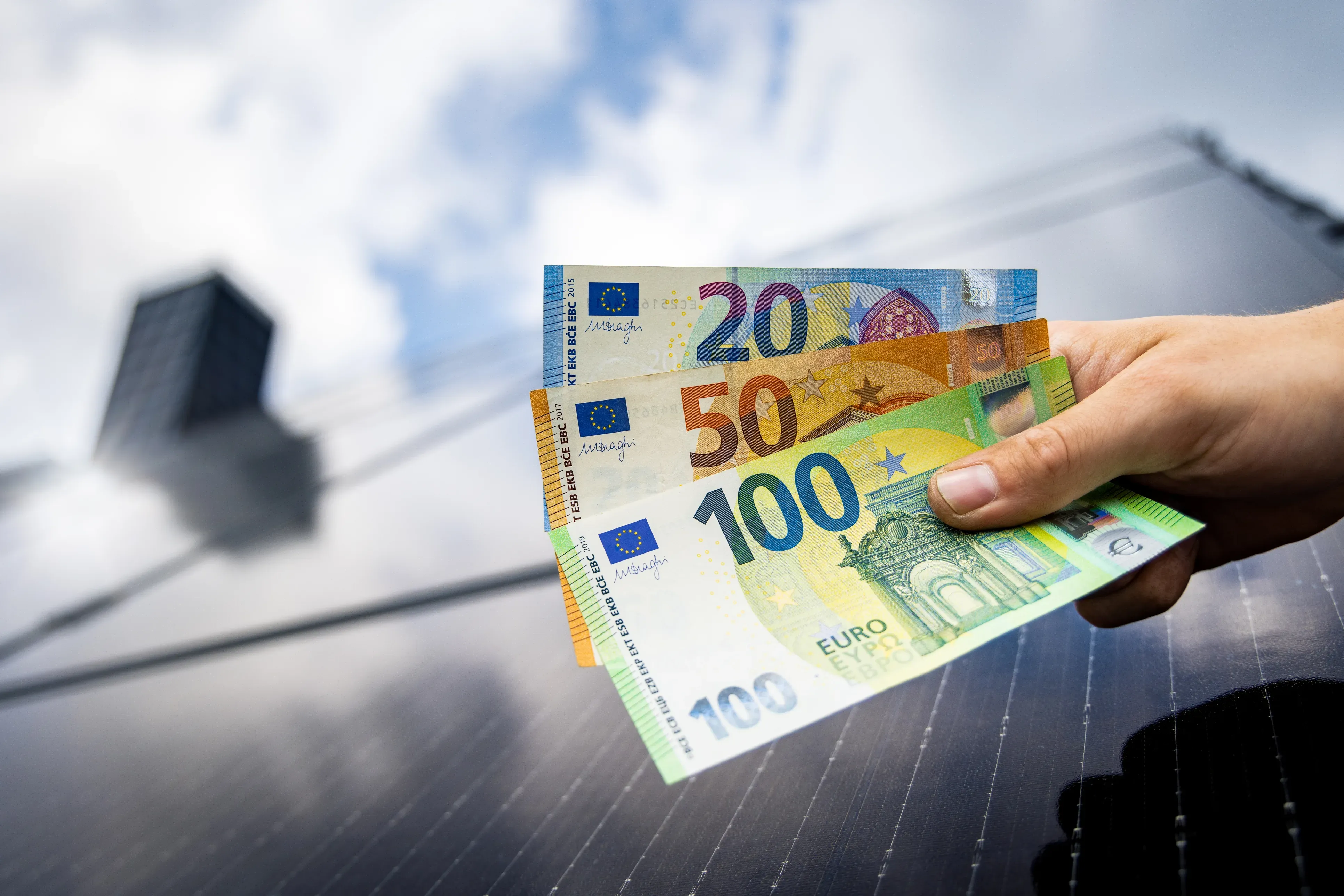Eine Hand hält mehrere Euro-Banknoten vor einem Solarmodul als Symbolbild für die Einspeisevergütung von Photovoltaikanlagen.