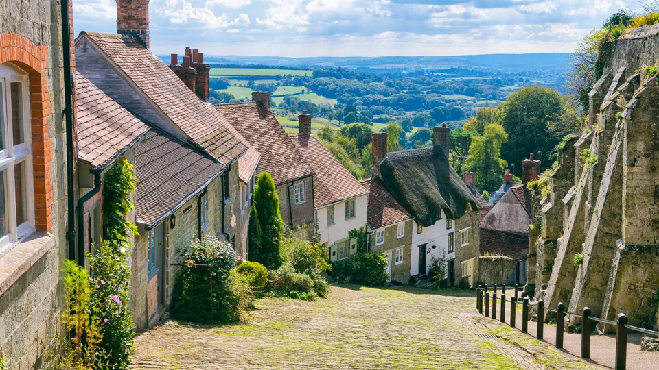 Cobblestone street lined with historic stone cottages overlooking rolling countryside.
