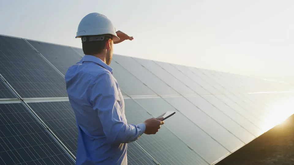 Arbeiter mit Schutzhelm inspiziert Solarmodule. Er schaut der Sonnen entgegen und hält ein Tablet in der Hand.