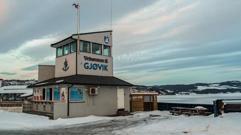 Liten bygning med skiltet «Velkommen til Gjøvik» ved en snødekt strandpromenade