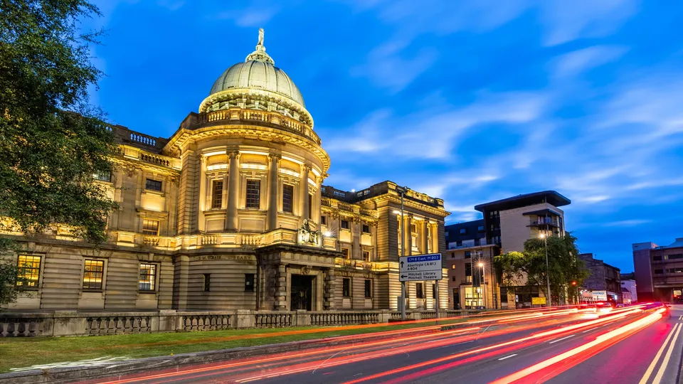 Historic stone building with golden dome illuminated at dusk, car light trails streaking across street.