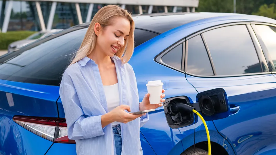Kvinde holder kaffe og smartphone mens hun oplader elektrisk køretøj.