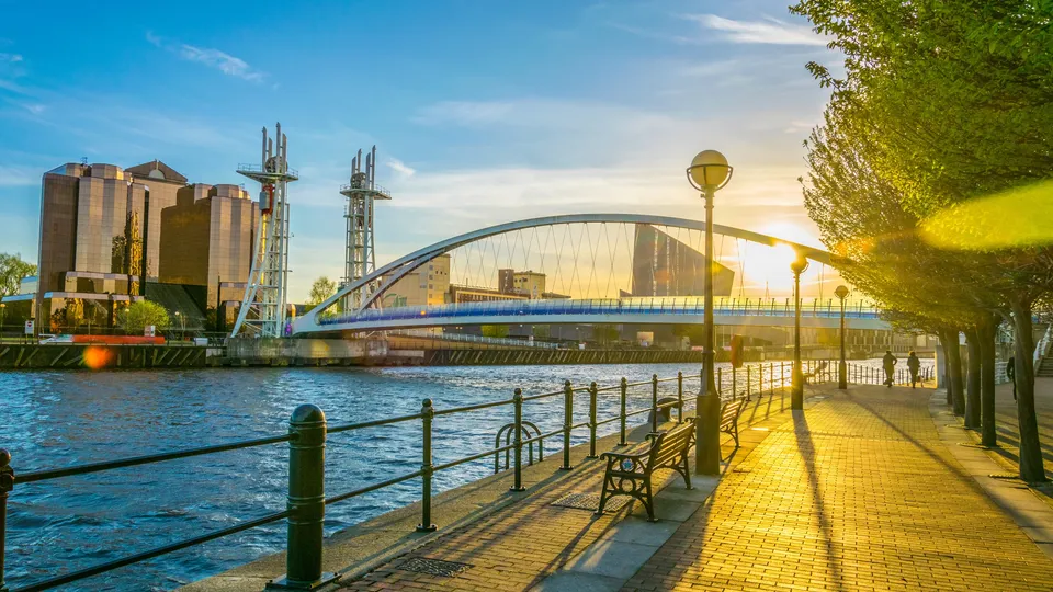 Waterfront promenade with benches overlooking a modern arched bridge and industrial buildings at sunset.