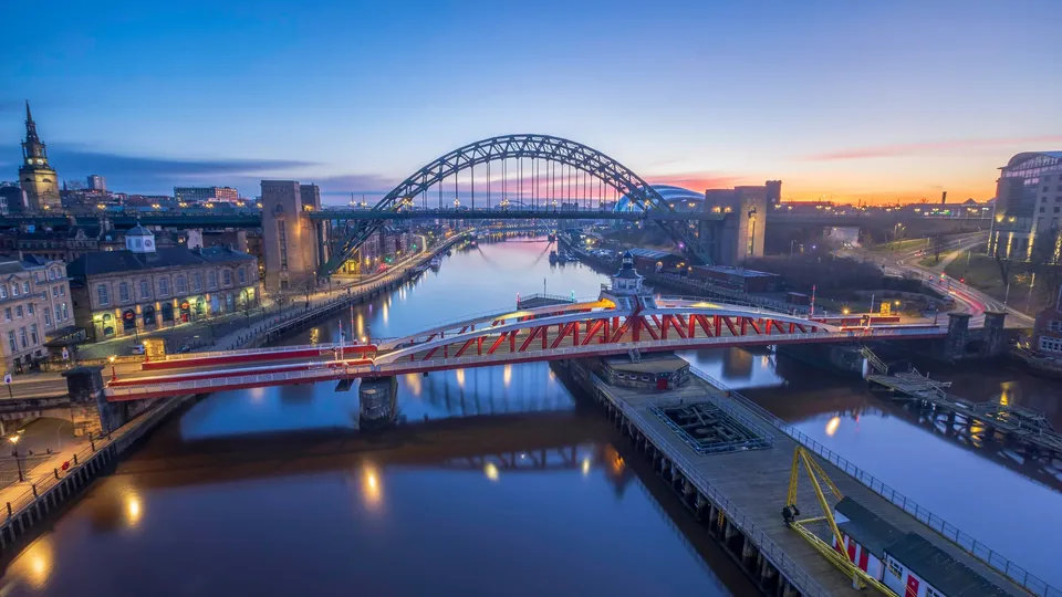 Newcastle cityscape at dusk with Tyne Bridge and red pedestrian bridge over river.