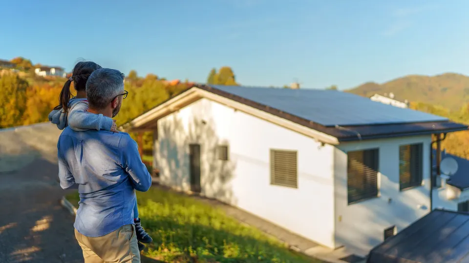 Mann hält Kind und blickt auf Haus mit Solarpanelen auf dem Dach.