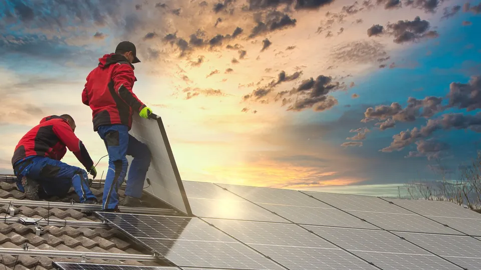 Two workers installing solar panels on a roof at sunset.