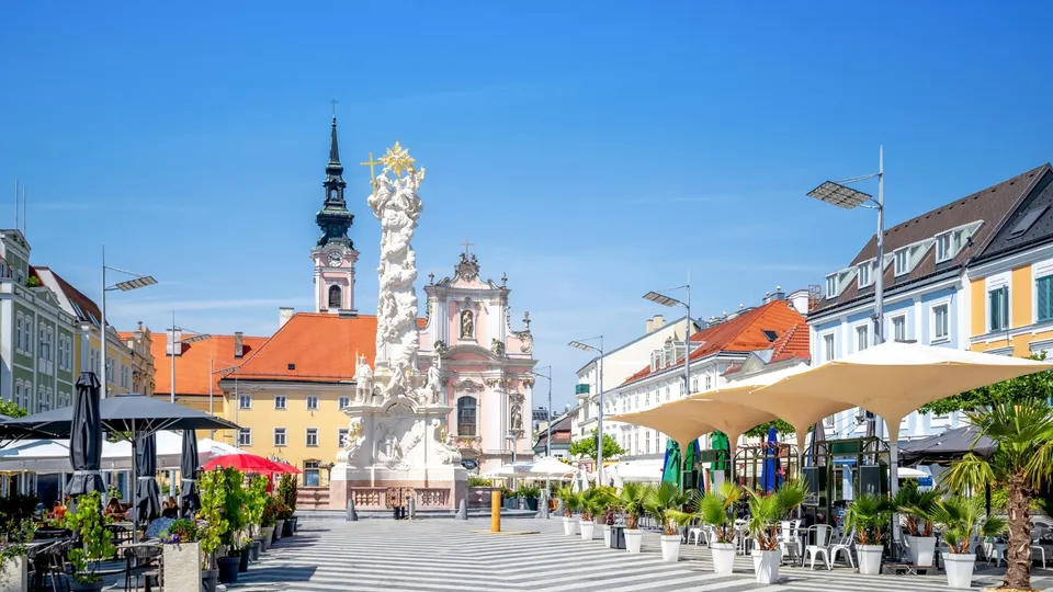 Marktplatz von St.Pölten mit Barockkirche, weißer Statue und farbigen Gebäuden unter blauem Himmel.