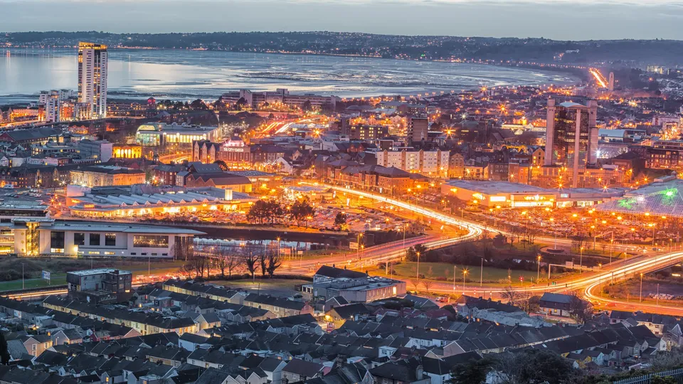 Illuminated cityscape at dusk overlooking a bay with residential neighborhoods in foreground.
