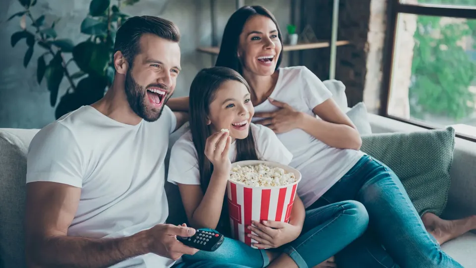 Three people laughing while watching television on a couch, one holding a popcorn bucket.