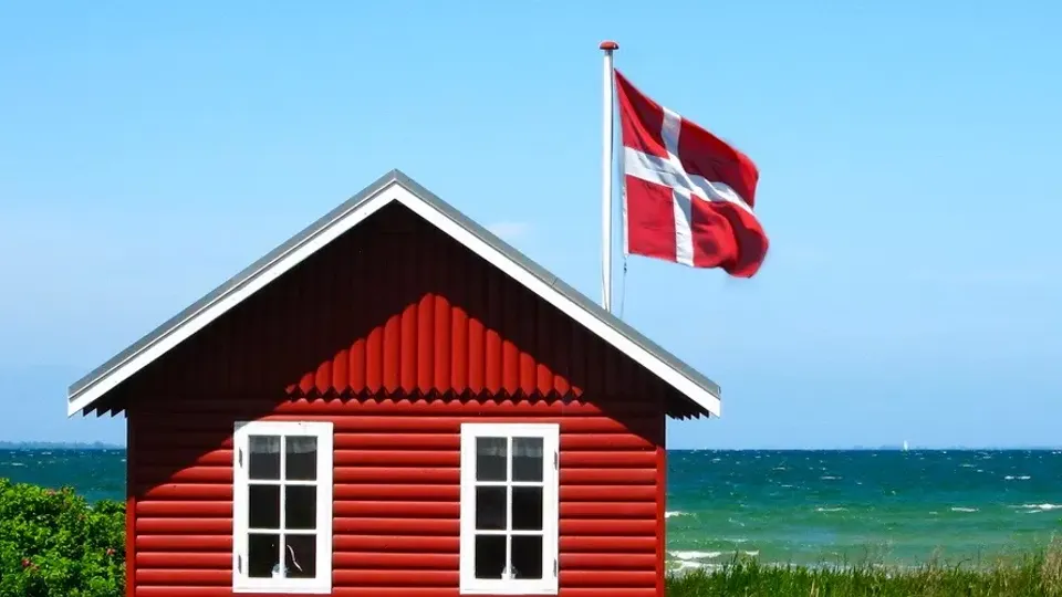 Et rødt sommerhus med hvide vinduer står ved en græsklædt strand under en klar blå himmel. Et dansk flag vajer på en stang og fremkalder et fredeligt kystsceneri.
