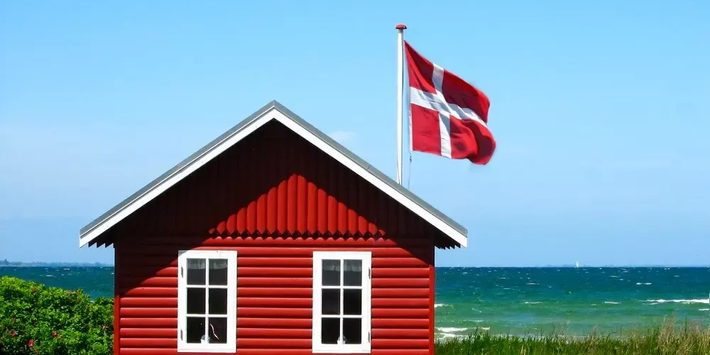 Et rødt sommerhus med hvide vinduer står ved en græsklædt strand under en klar blå himmel. Et dansk flag vajer på en stang og fremkalder et fredeligt kystsceneri.