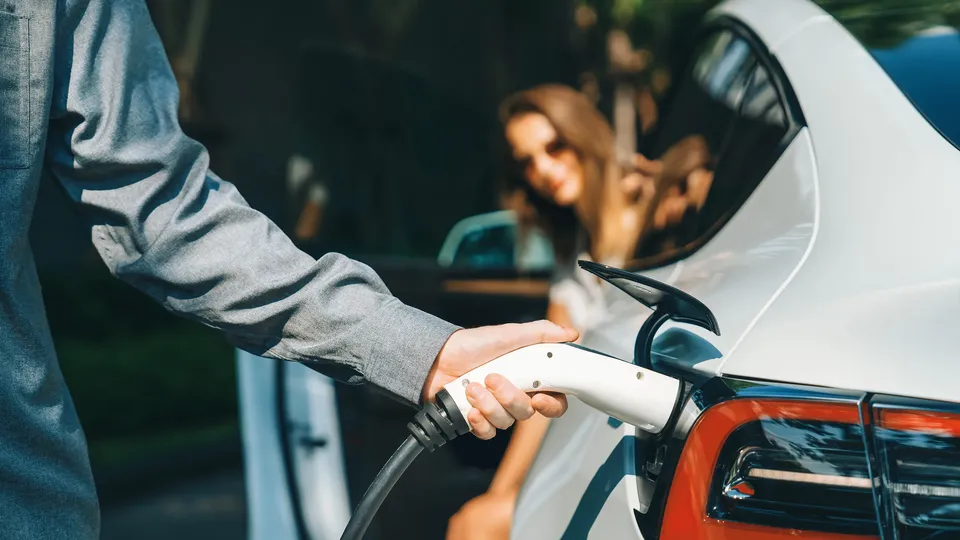 A person charging their electric vehicle with a person sat in the car in the background.
