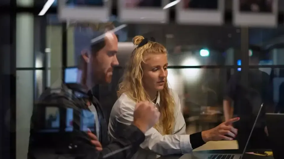 Nettbureau-ansat peger på laptopskærm mens en kollega ser til på kontoret.