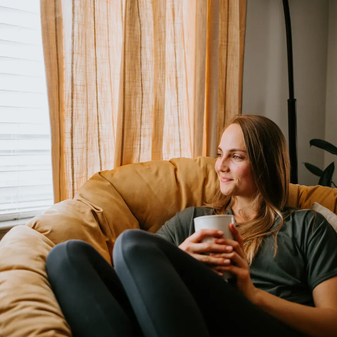 Mujer sentada en el sofá sosteniendo una taza, mirando hacia la ventana.