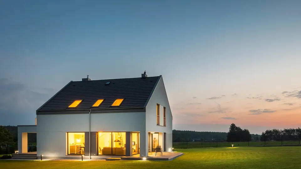 Casa moderna blanca con ventanas iluminadas al atardecer en un paisaje rural.