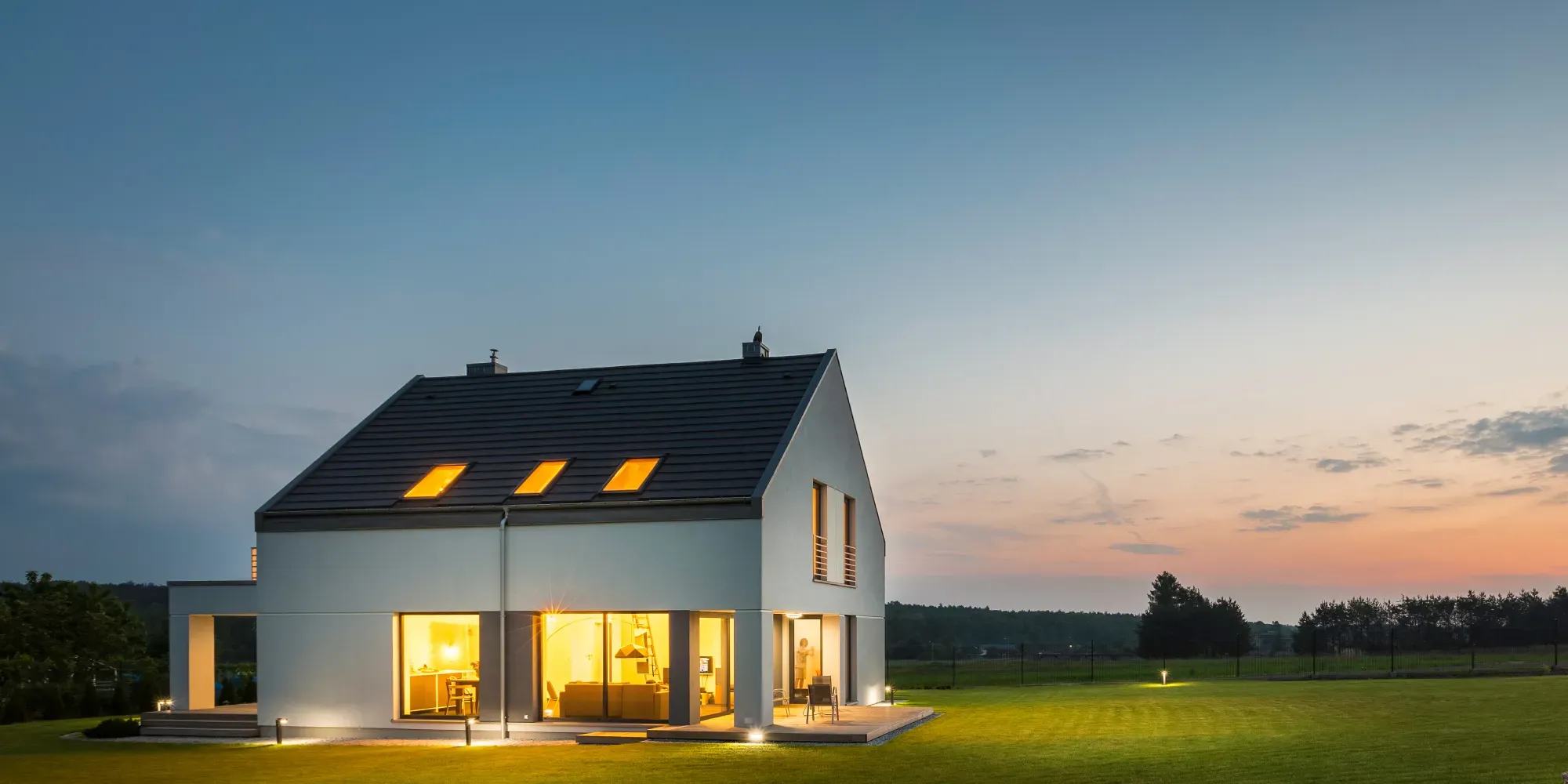 Casa moderna blanca con ventanas iluminadas al atardecer en un paisaje rural.