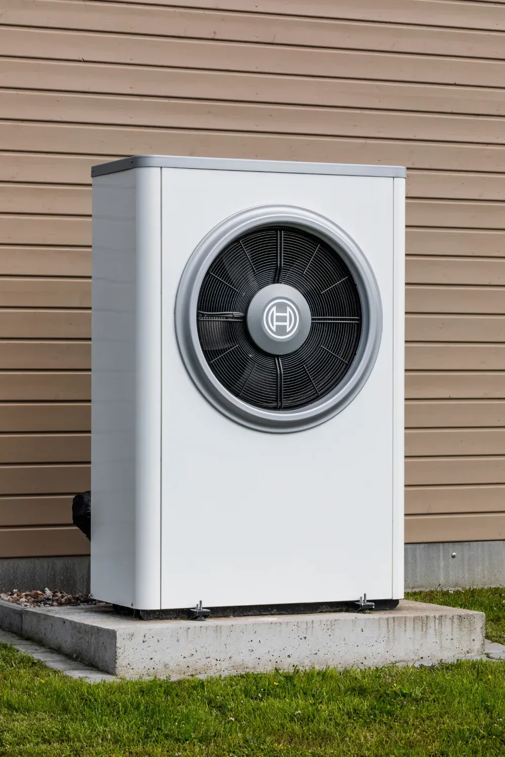 White heat pump unit with large black fan mounted on concrete pad against wooden siding.