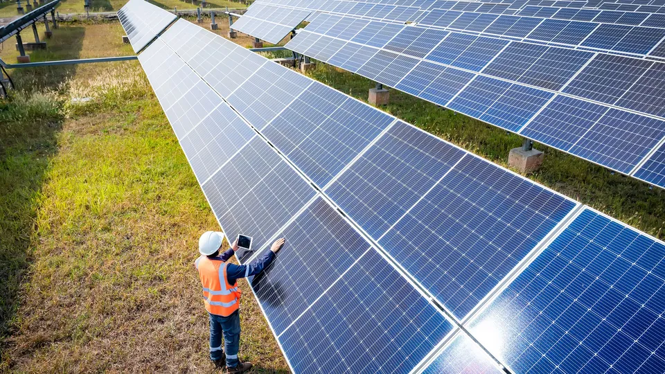 Solar farm technician in orange safety vest and white hard hat inspecting photovoltaic panels with a tablet.