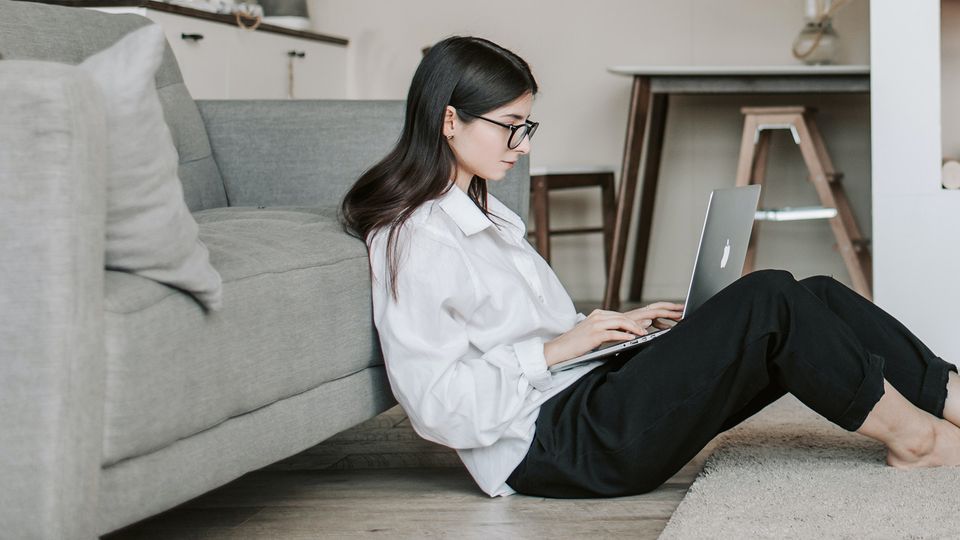 Bilde av en kvinne som sitter på gulvet inntil en sofa og skriver på en laptop.