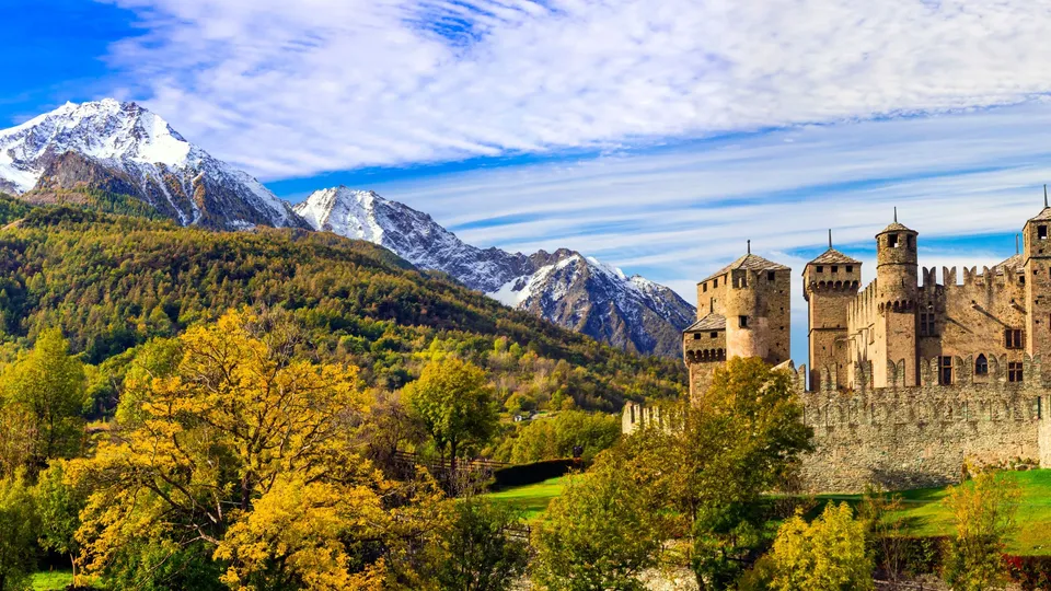 Castello medievale con torri che domina una valle boscosa e montagne innevate.