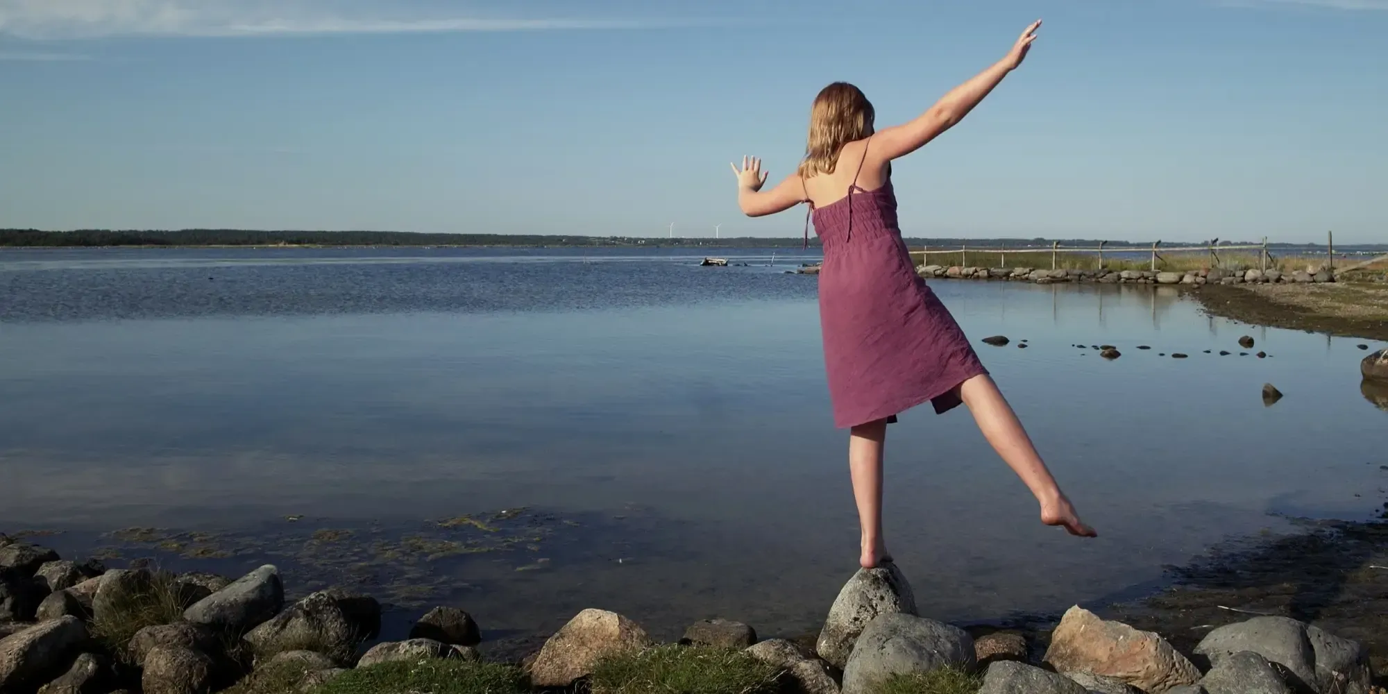 En flicka balanserar på stenar med armarna utsträckta vid vattnet.