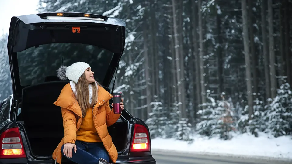 Kvinde sidder i bagagerummet af en åben bil og holder en kop i hånden i en snedækket skov.