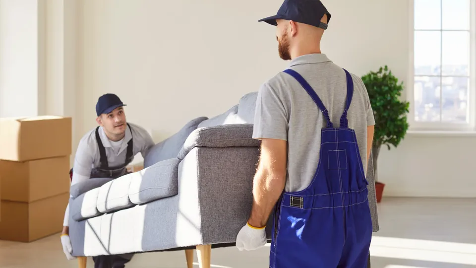 To flyttearbeidere bærer en sofa i et tomt rom.