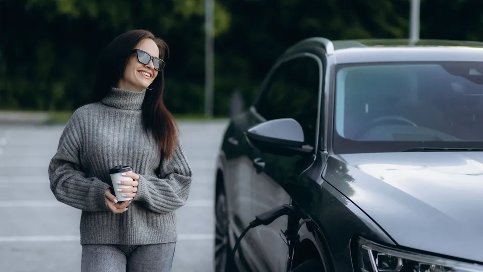 Kvinde med kaffekop står ved siden af elbil under opladning.