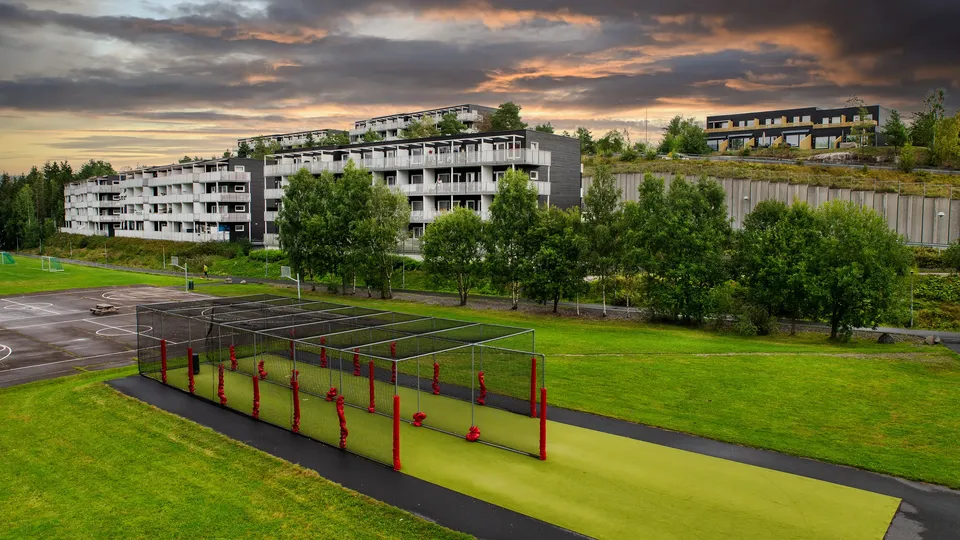 Moderne leilighetsblokker med utsikt over idrettsanlegg og grønt område ved solnedgang.