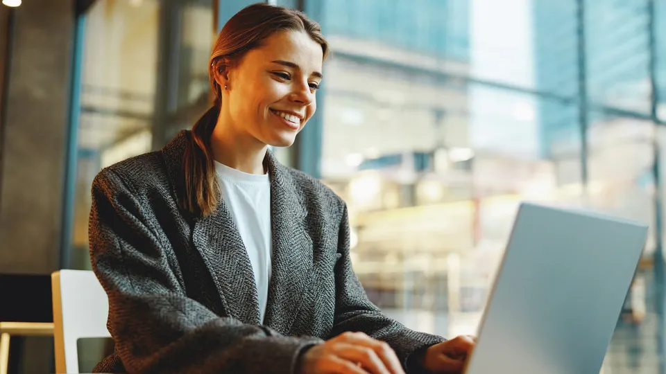Kvinde smiler mens hun skriver på bærbar computer ved skrivebord ved vindue.