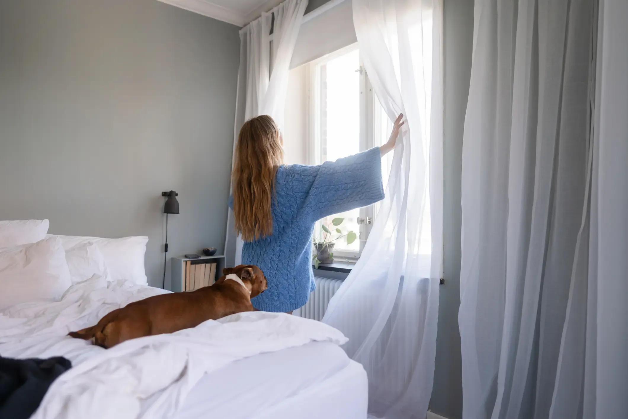 Kvinna öppnar gardiner medan hund sträcker sig på sängen.