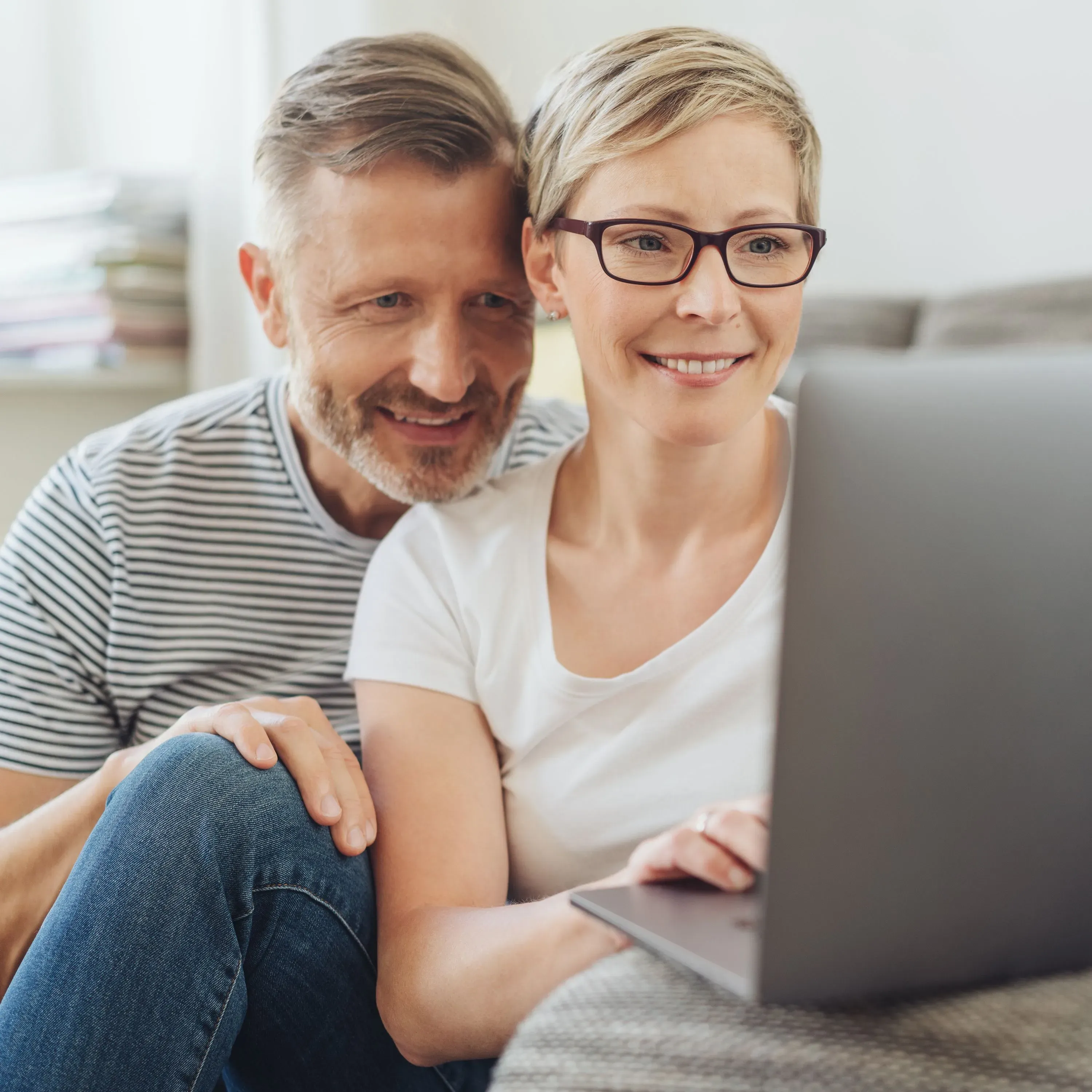 Ein Pärchen im mittleren Alter sitzt gemeinsam auf dem Boden und schaut auf einen Laptop-Bildschirm. Sie vergleichen Immobilienmakler online.