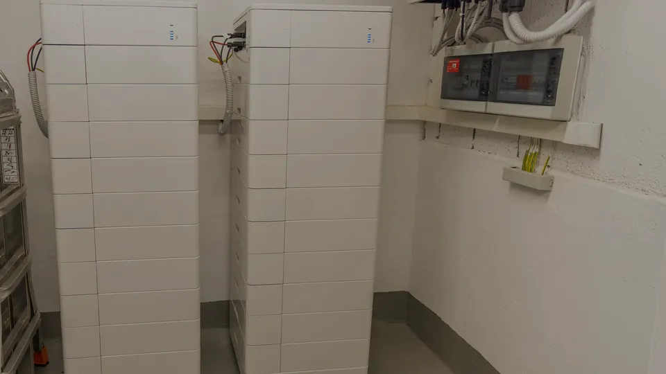 Två höga vita hembatterier som står sida vid sida i ett teknikrum, anslutna till en elcentral på väggen.
