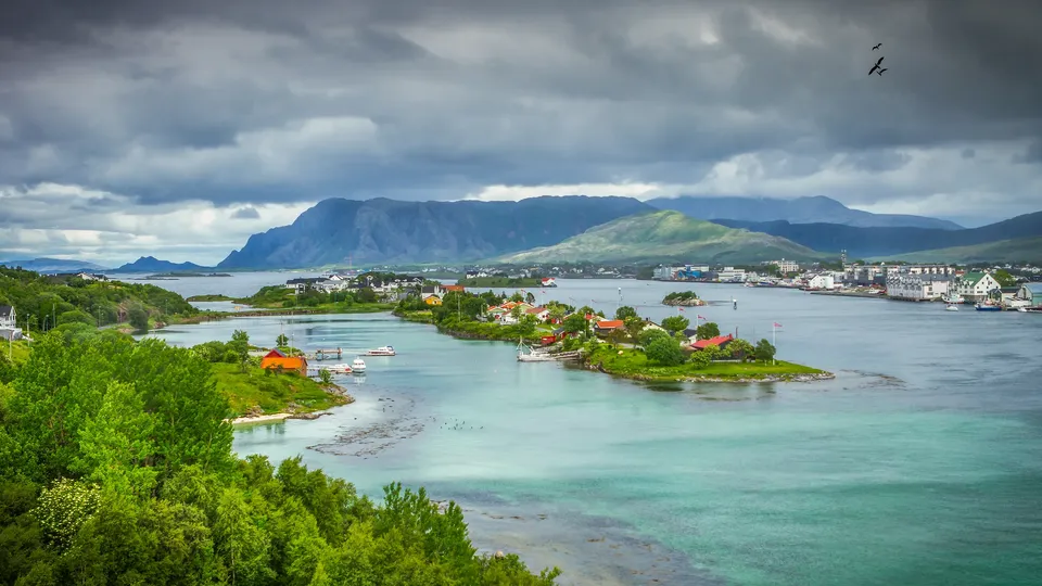 Kystbebyggelse på øyer omgitt av turkisblått vann og fjell under skydekke.