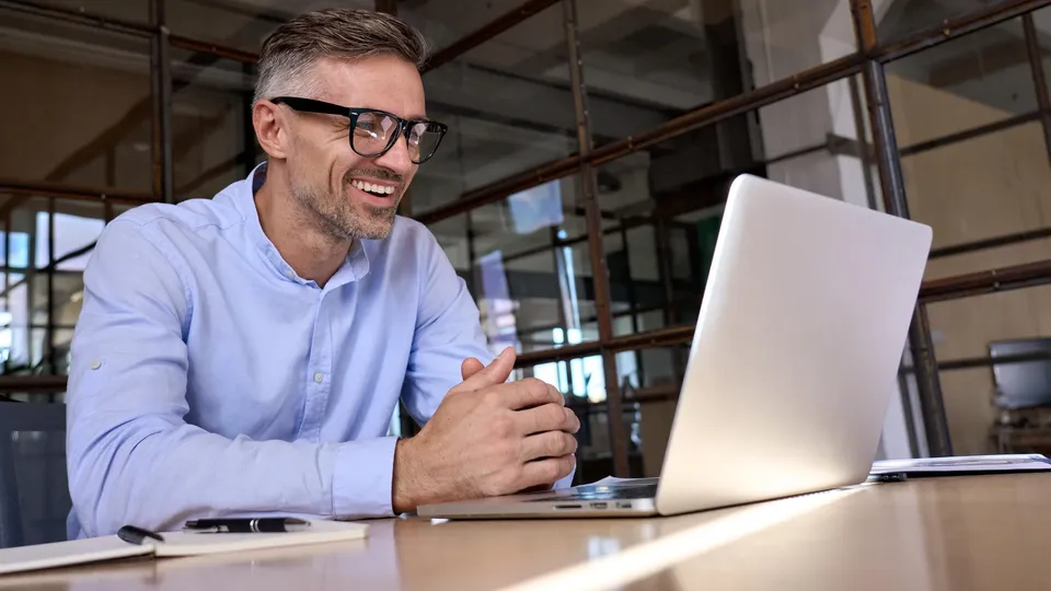 Lächelnder Geschäftsmann mit Brille, der im Büro an einem Laptop arbeitet.