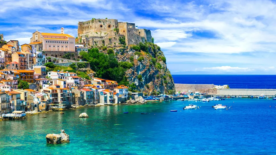 Villaggio costiero colorato con rovine di castello su scogliera rocciosa che si affaccia sul mare turchese.