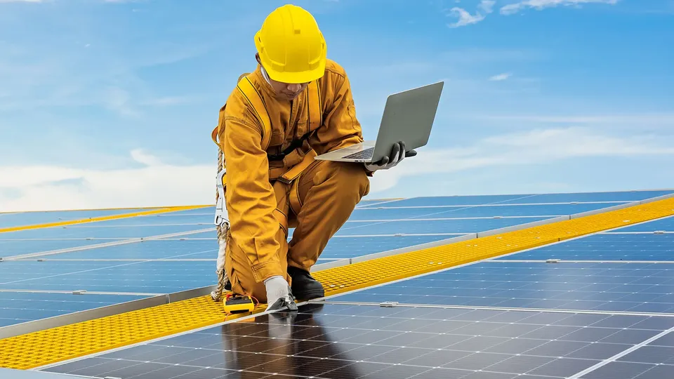 Construction worker in safety gear using laptop on solar panel installation.