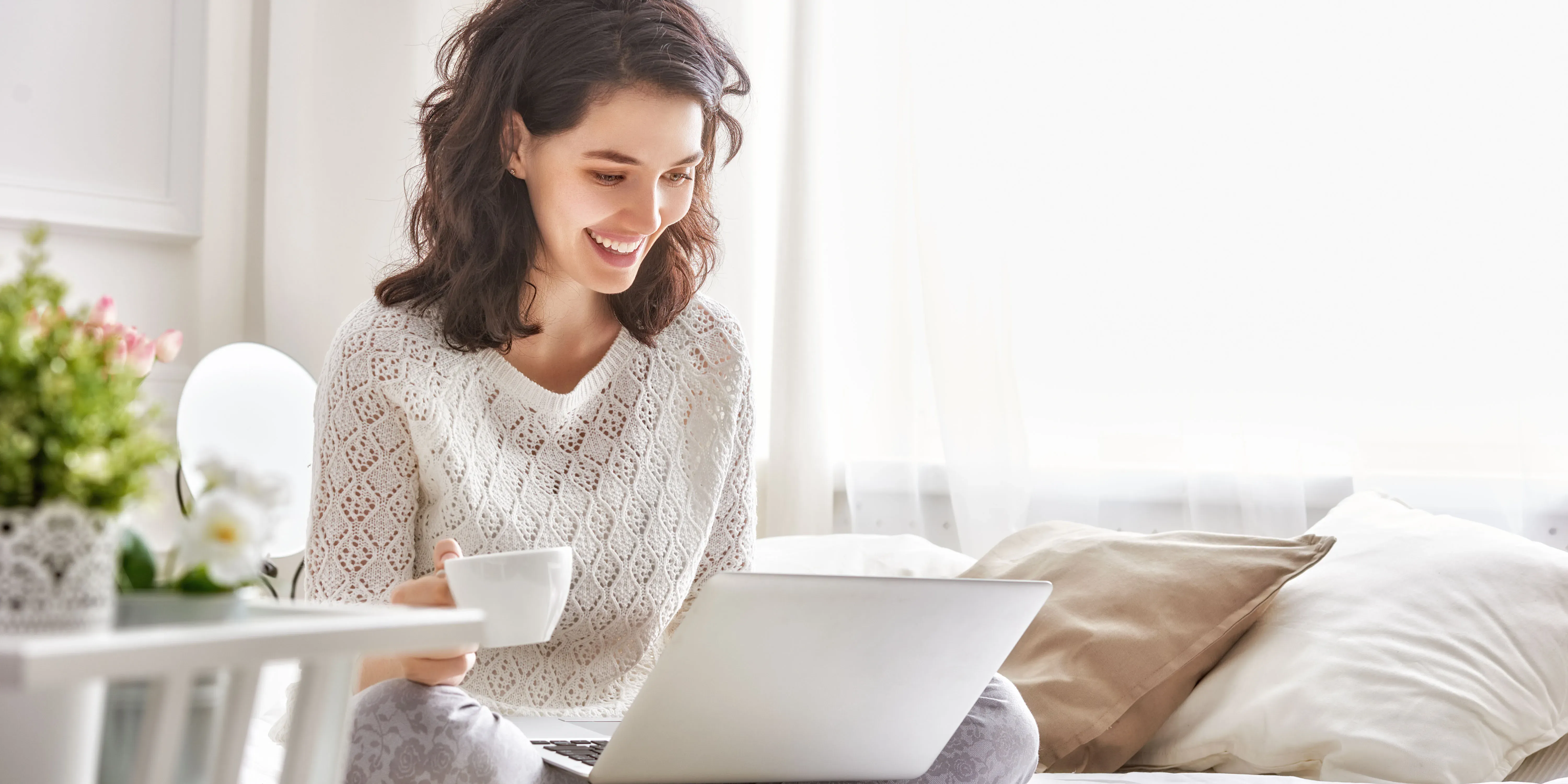 Mujer sonriendo mientras usa laptop y sostiene taza de café.