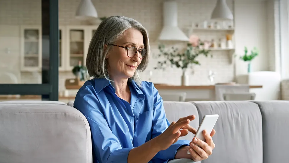 Mujer de mediana edad con cabello gris y lentes sentada en un sofá usando un smartphone.