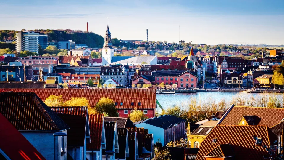Farverig byfront med kirketårne og røde tage ved en havnefront.