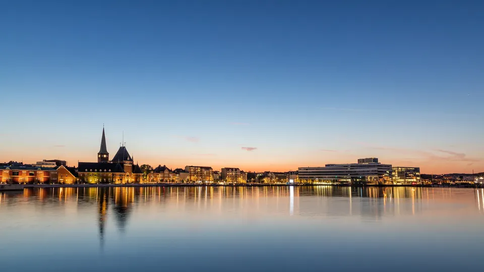 Bylandskab ved vandet i skumringen med oplyst bygninger reflekteret i stille vand.