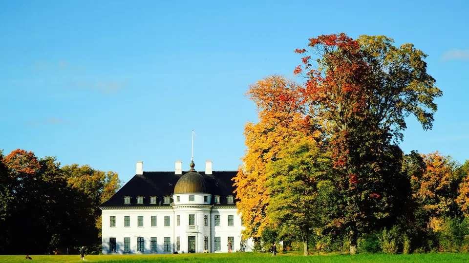 Hvidt herskabsbolig med kuppel omgivet af efterårstræer og grøn græsplæne.