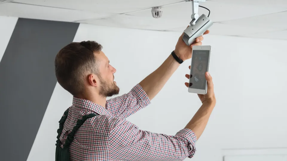 Hombre sosteniendo un smartphone y un dispositivo mientras ajusta un sistema de seguridad montado en el techo.