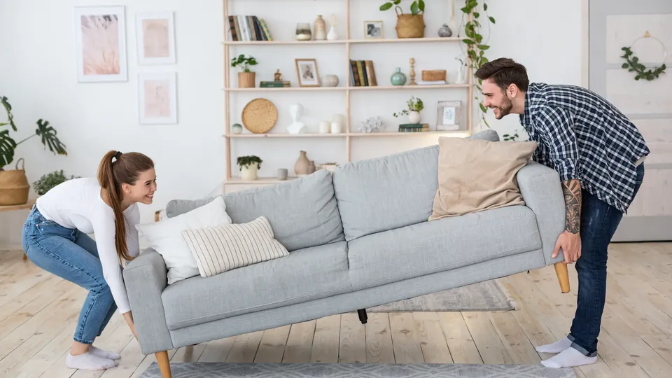 Par løfter og flytter en grå sofa sammen.