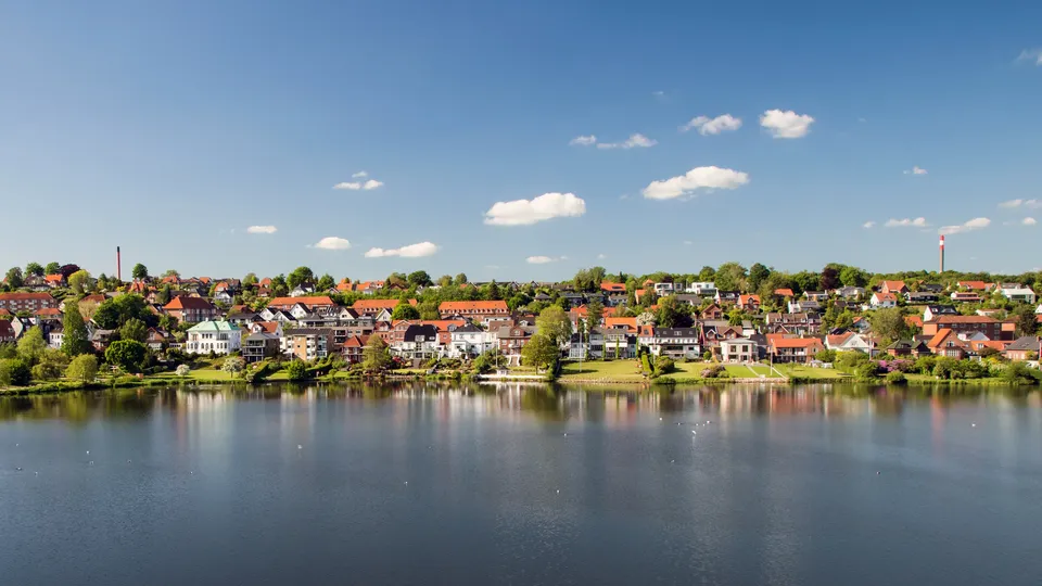 Billede af Koldning: By ud til fjorden med huse, der har røde tage reflekteret i stille vand under blå himmel.